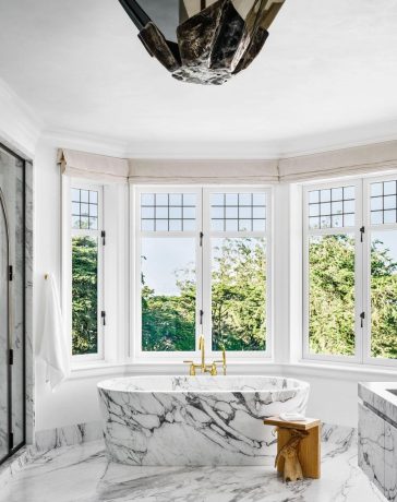 rounded bathroom with double windows in an arc at the back with a standalone marble tub at center and a floating marble vanity and a large bat like light fixture at center and a glass shower at left