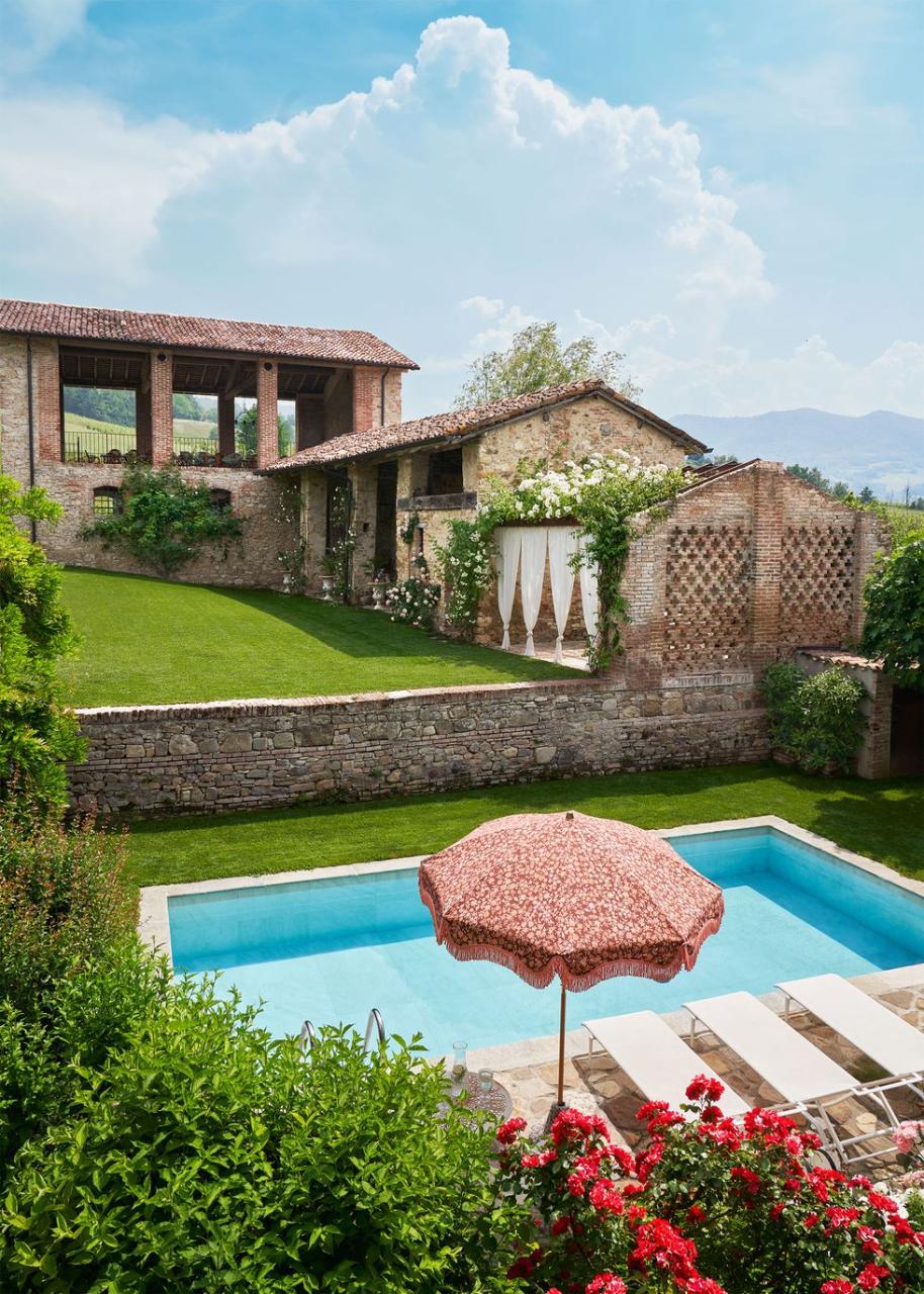 12th century buildings with open windows lead to a rectangular pool with chaises longue, an umbrella, shrubs and flowering bushes, and a stone retainer wall next to a grassy lawn