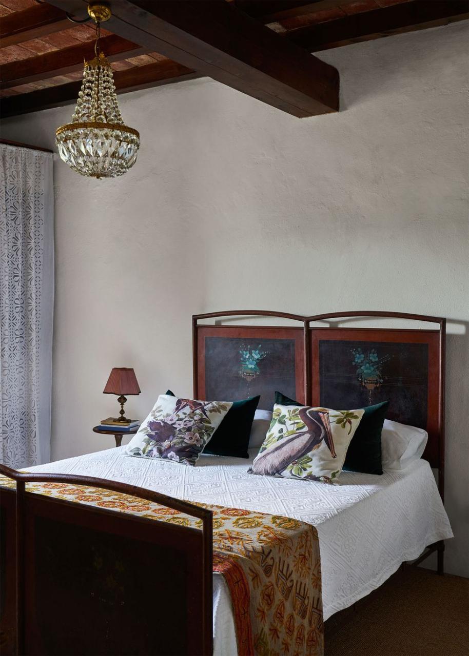 a metal bed with flowers painted on headboard, white bedspread with printed accent pillows and embroidered throw, small nightstand with lamp, glass chandelier, plaster walls, and beamed ceiling