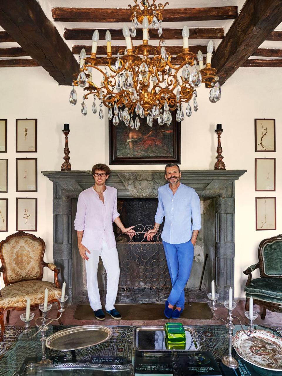 two men pose for a picture in front of a fireplace with a filigree screen, two antique chairs, artworks above and beside fireplace, two candlesticks on mantel, cocktail table with candelabras and trays, glass chandelier