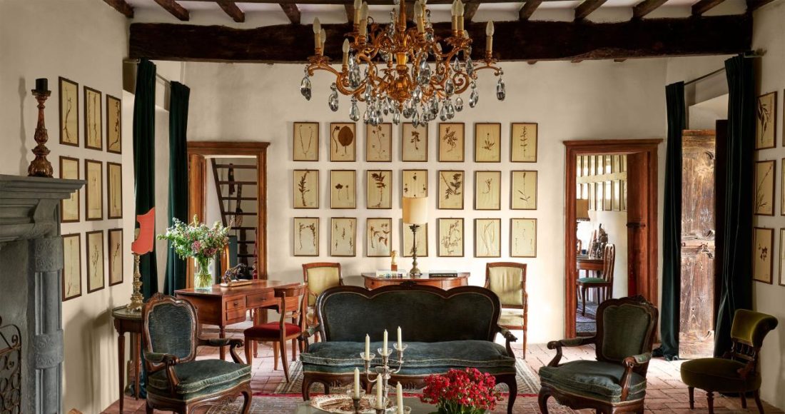 a room with a crystal chandelier, vintage sofa and chairs with velvet cushions, cocktail table with candelabras and trays, oriental rug, wooden desk and chair, a wall with 21 small framed botanical art pieces, beamed ceiling