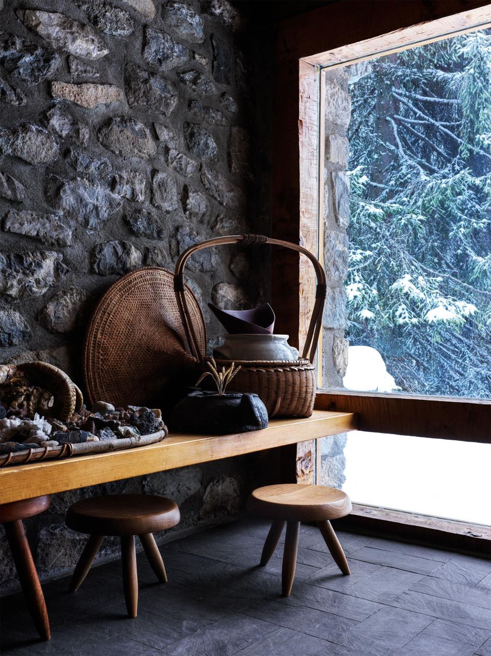 a table with a basket and a other items on it and three legged stools and stone walls and a view looking out to a snowy landscape