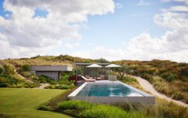 by a dirt road is a swimming pool with umbrellas and chairs by a pool house built into the grassy hillside