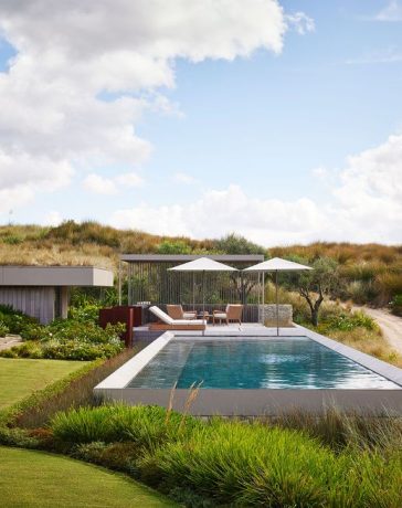 by a dirt road is a swimming pool with umbrellas and chairs by a pool house built into the grassy hillside