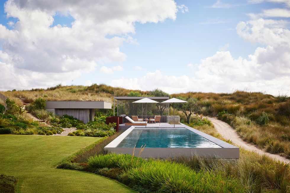 by a dirt road is a swimming pool with umbrellas and chairs by a pool house built into the grassy hillside