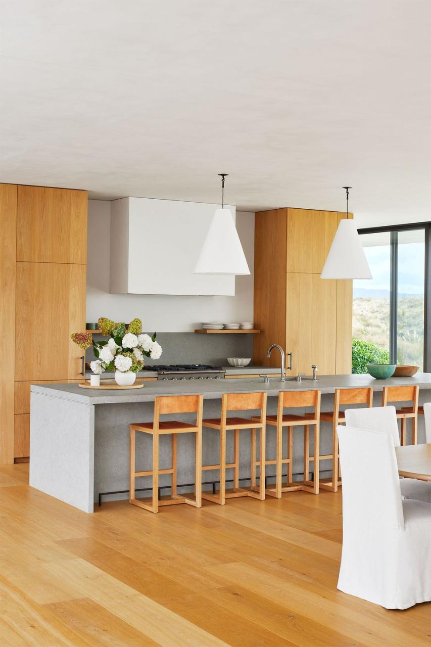 a kitchen with a wood floor and wood floor to ceiling cabinets, gray limestone island with wood stools, two large cone shaped white pendants, glass wall