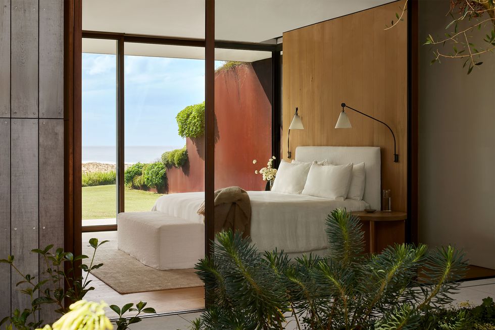 a room with glass walls has a bed with fabric headboard against a wood panel with long armed sconces, white bedding, a foot bench, and a view of the grass lawn and sea beyond