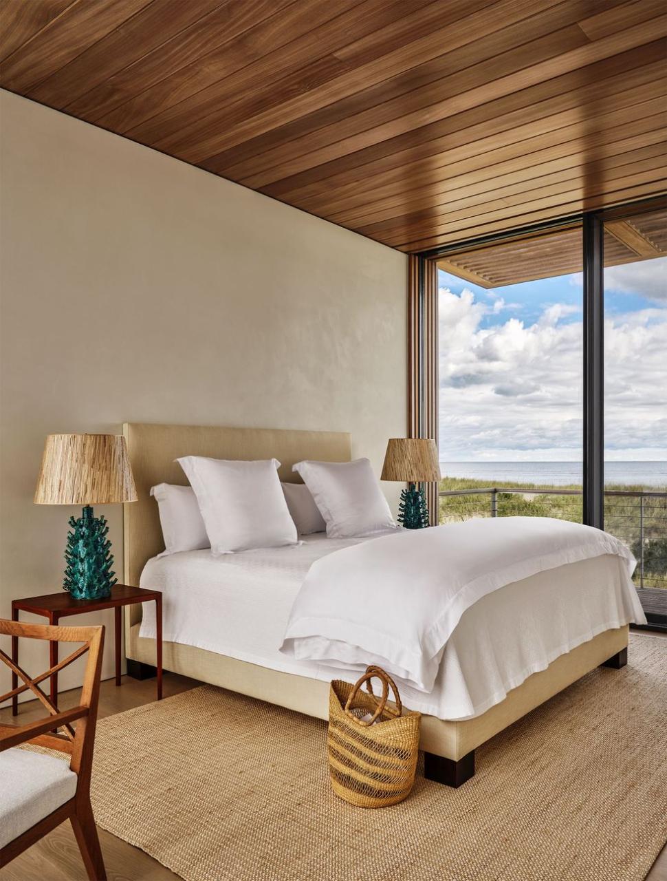 a bed with upholstered headboard and frame, white bedding, nightstands with lamps with bases resembling coral, natural rug, basket by bed, wood ceiling and floor, glass doors to terrace above the dunes