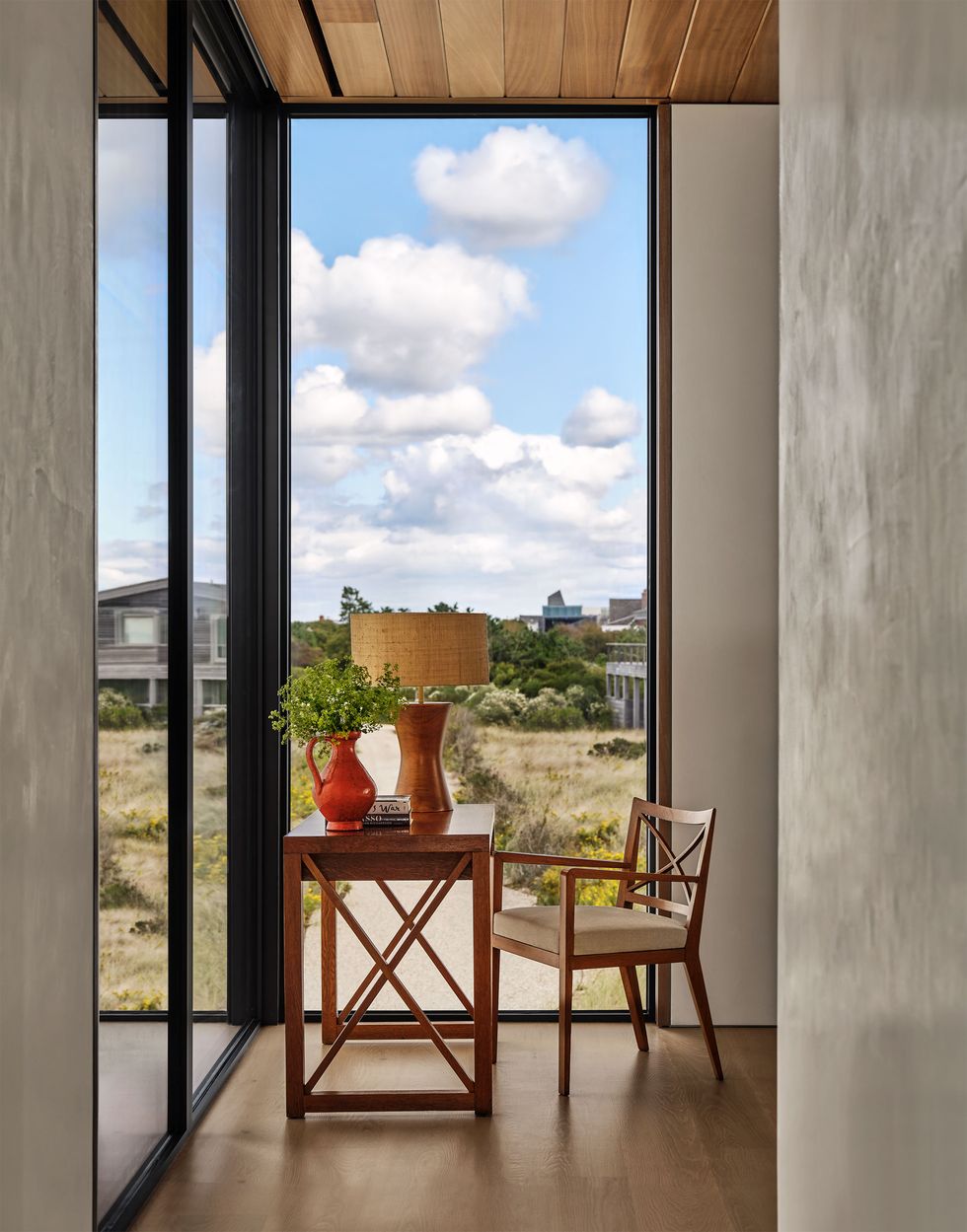 a corner of a room with a small desk and chair, a lamp, vase, and books on top of desk, plaster interior walls and two glass walls looking out to grassy dunes and other homes