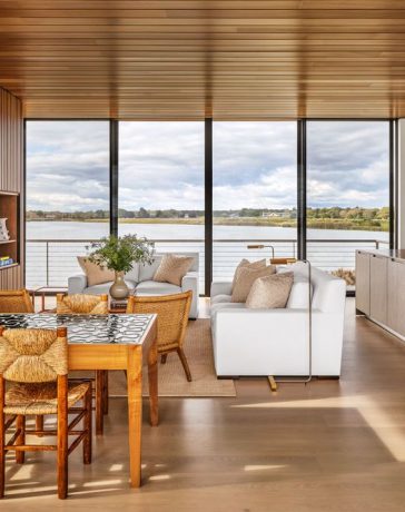 a large open room with a glass wall looking out to the sea, dining table and chairs, sofa, loveseat, and cocktail table, slatted wood wall with inset shelves, kitchen island with sink and cabinets, wood ceiling and floor