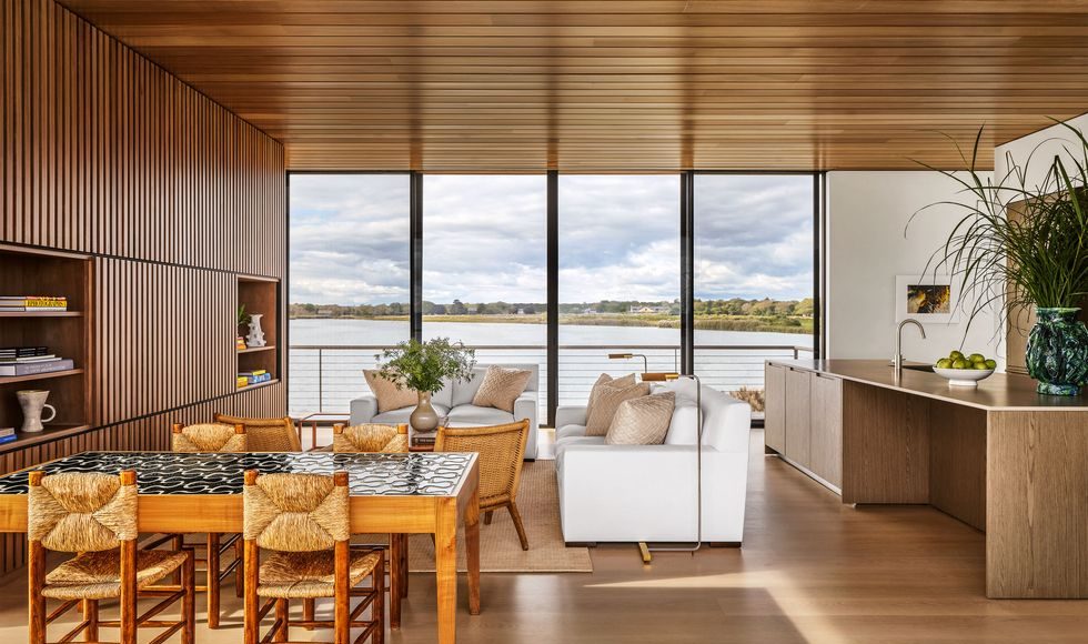 a large open room with a glass wall looking out to the sea, dining table and chairs, sofa, loveseat, and cocktail table, slatted wood wall with inset shelves, kitchen island with sink and cabinets, wood ceiling and floor