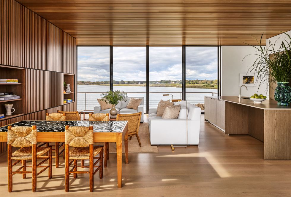 a large open room with a glass wall looking out to the sea, dining table and chairs, sofa, loveseat, and cocktail table, slatted wood wall with inset shelves, kitchen island with sink and cabinets, wood ceiling and floor