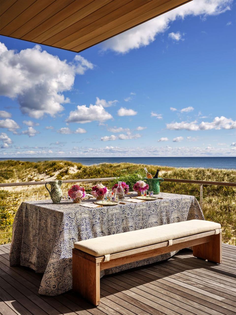 a deck a wood and wired railing looking out over grassy dunes and the sea, table set with dishes and flowers, printed tablecloth, bench with cushion seat