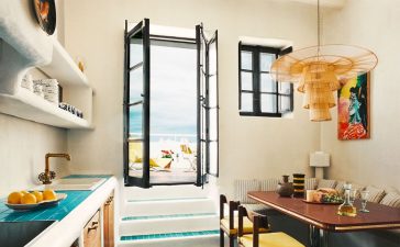 a kitchen with turquoise tiled floor and a wood table with yellow chairs and a banquette with striped fabric, rattan pendant, counter with sink and shelves above and wood cabinets below, steps go to a doorway out to terrace