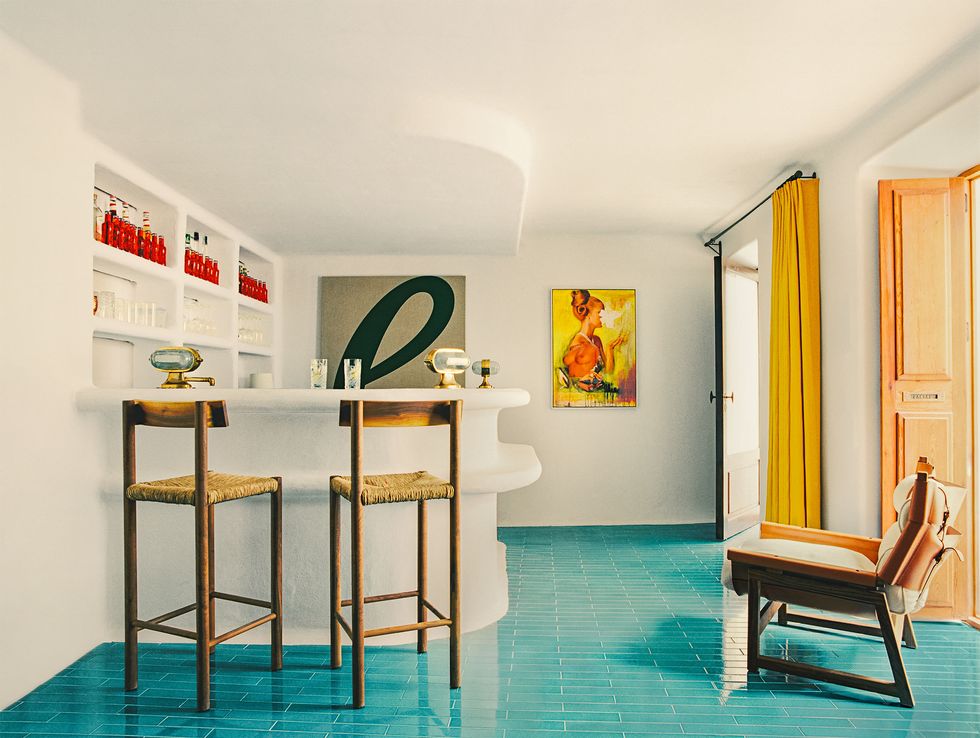 a room with white walls has a white stone bar with bottles inset in wall above, curved counter and and stools with rush seats, turquoise colored tile floors and yellow drapes and a door leading outside