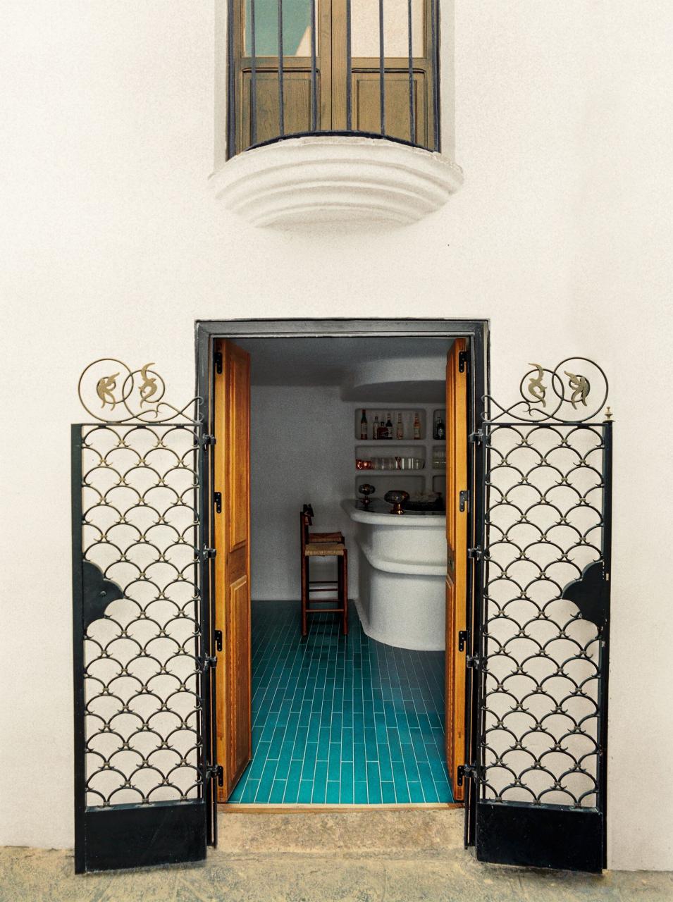 outside doorway with wrought iron exterior doors and wooden interior doors, inside is a turquoise tiled floor, white walls and a curved stone bar with barstools and bottles inset in wall above bar