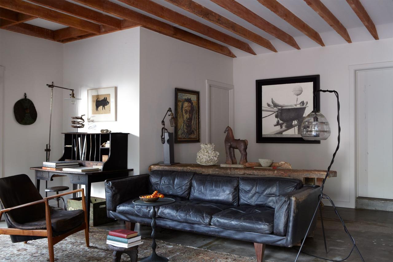 white painted living room with wood beam ceiling and a black leather sofa at center and artwork in black frames on walls and a desk in the alcove corner by the window