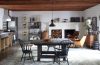 wood beam ceiling living dining space with a white brick fireplace at far end and a wood insert next to it and some modern wood club chairs and a dark square farmhouse table and chairs in the foreground and a kitchen set up to the left