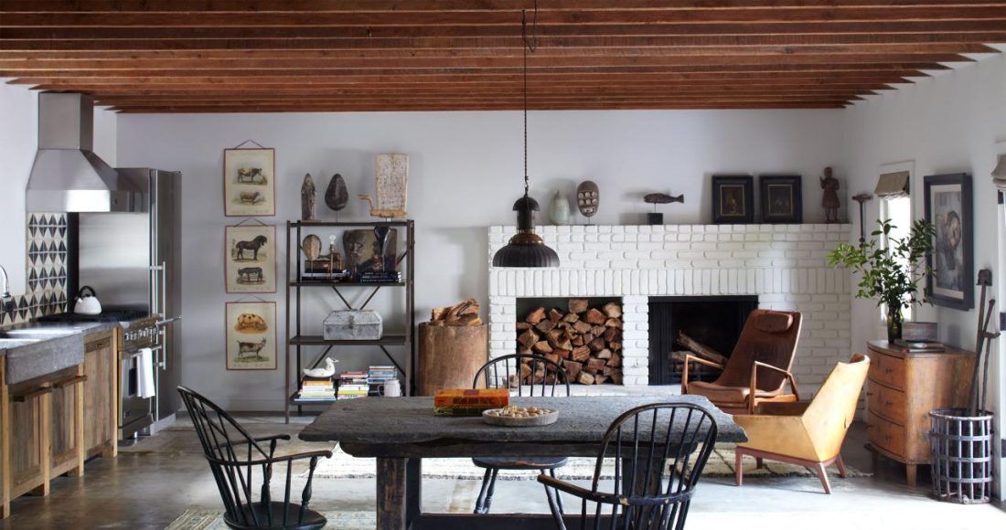 wood beam ceiling living dining space with a white brick fireplace at far end and a wood insert next to it and some modern wood club chairs and a dark square farmhouse table and chairs in the foreground and a kitchen set up to the left