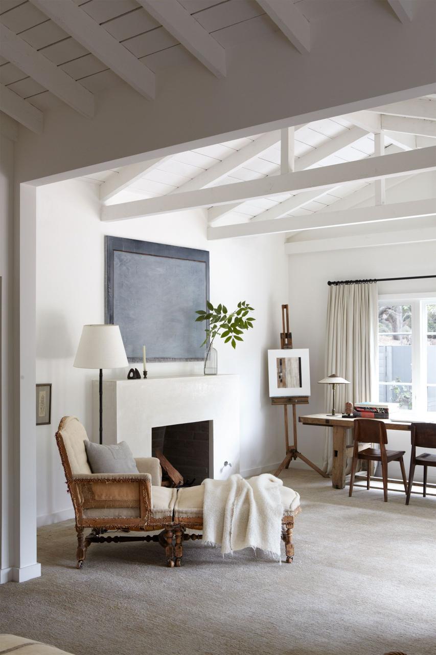 white room with beam ceilings and white fireplace and slate gray and blue artwork over it and a old two piece chaise with white throw over it and in the background a rustic table with some black chairs pulled up to it and an easel in the corner