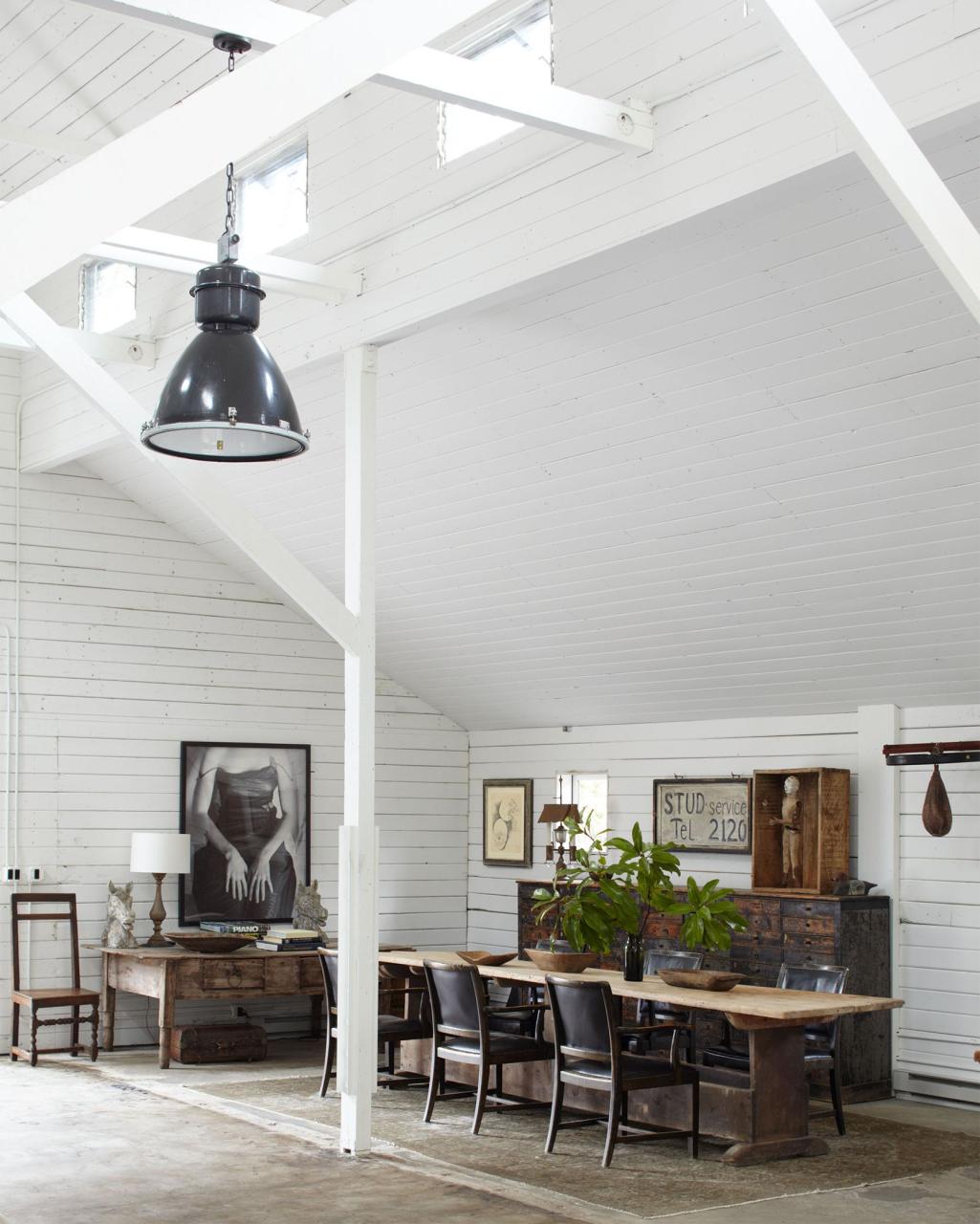 high ceilinged white barn room with long swedish table and black chairs pulled up to it on a thin rug and a rustic wood console with pigeon hold draws and a desk and artwork on the wood plant walls