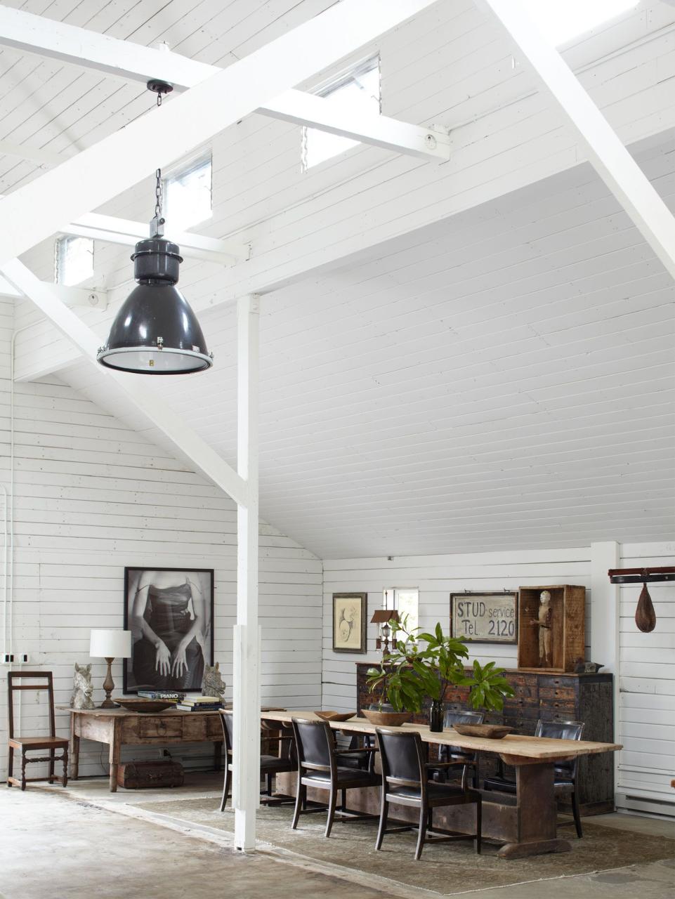 high ceilinged white barn room with long swedish table and black chairs pulled up to it on a thin rug and a rustic wood console with pigeon hold draws and a desk and artwork on the wood plant walls