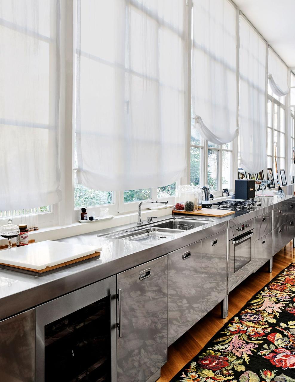 stainless steel kitchen cabinetry with multiple windows with white shades behind it and a dark flora rug on the floor