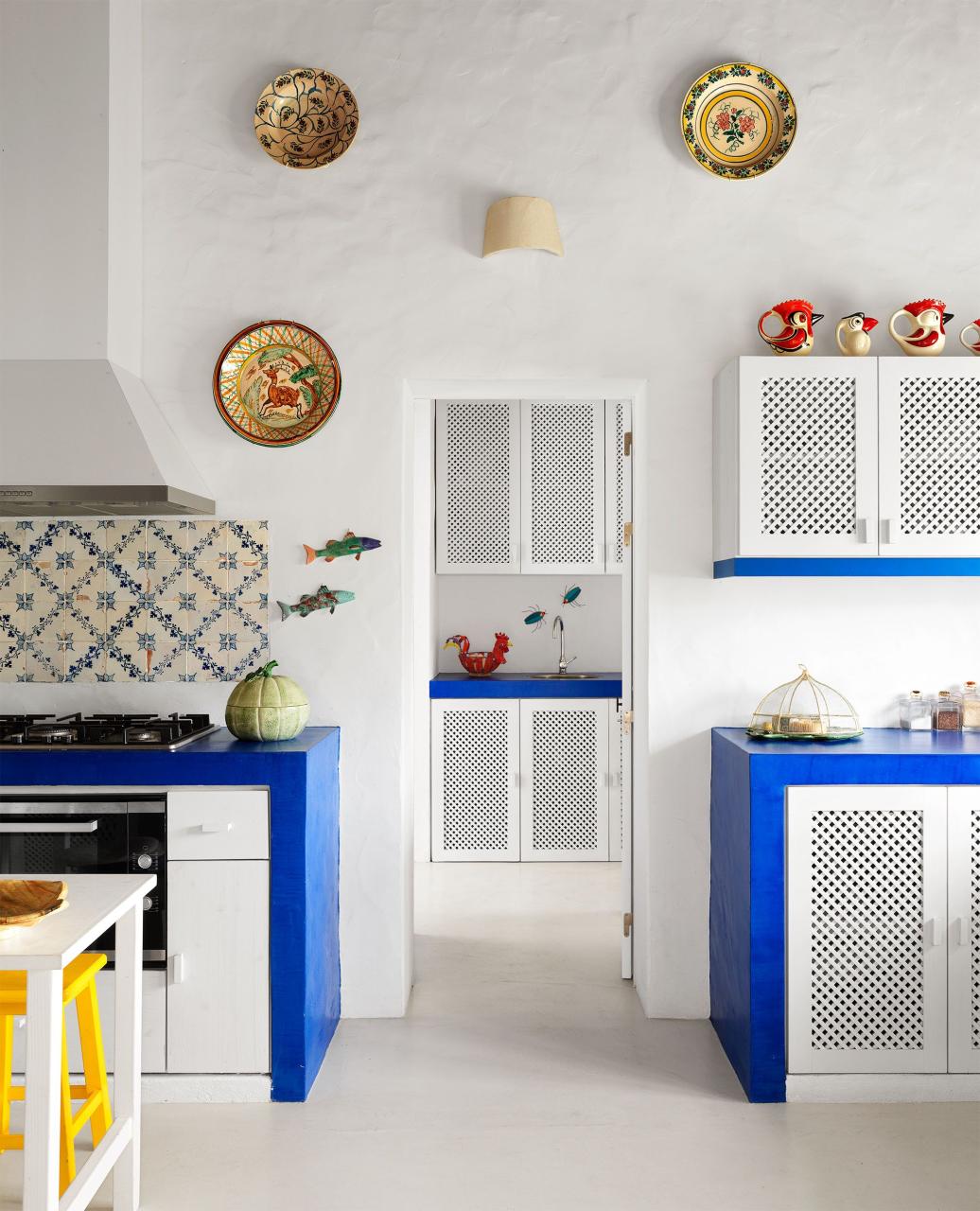 a kitchen with white walls and cabinets accented with deep blue counters, blue and white tiles, ceramic fish and portuguese plates adorn the walls, four ceramic pitchers in the shape of chickens top a cabinet