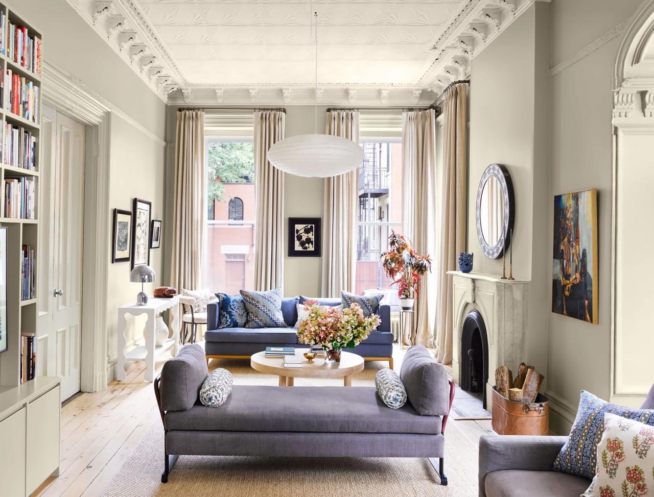 high ceilinged room with tall curtained windows at the back and plank floors and a fireplace at left against cream walls and a pale purple settee and sofa in pale blue with a round table with flowers at center and a oval hanging pendant lamp from pressed tin ceilings surrounded by intricate deep molding