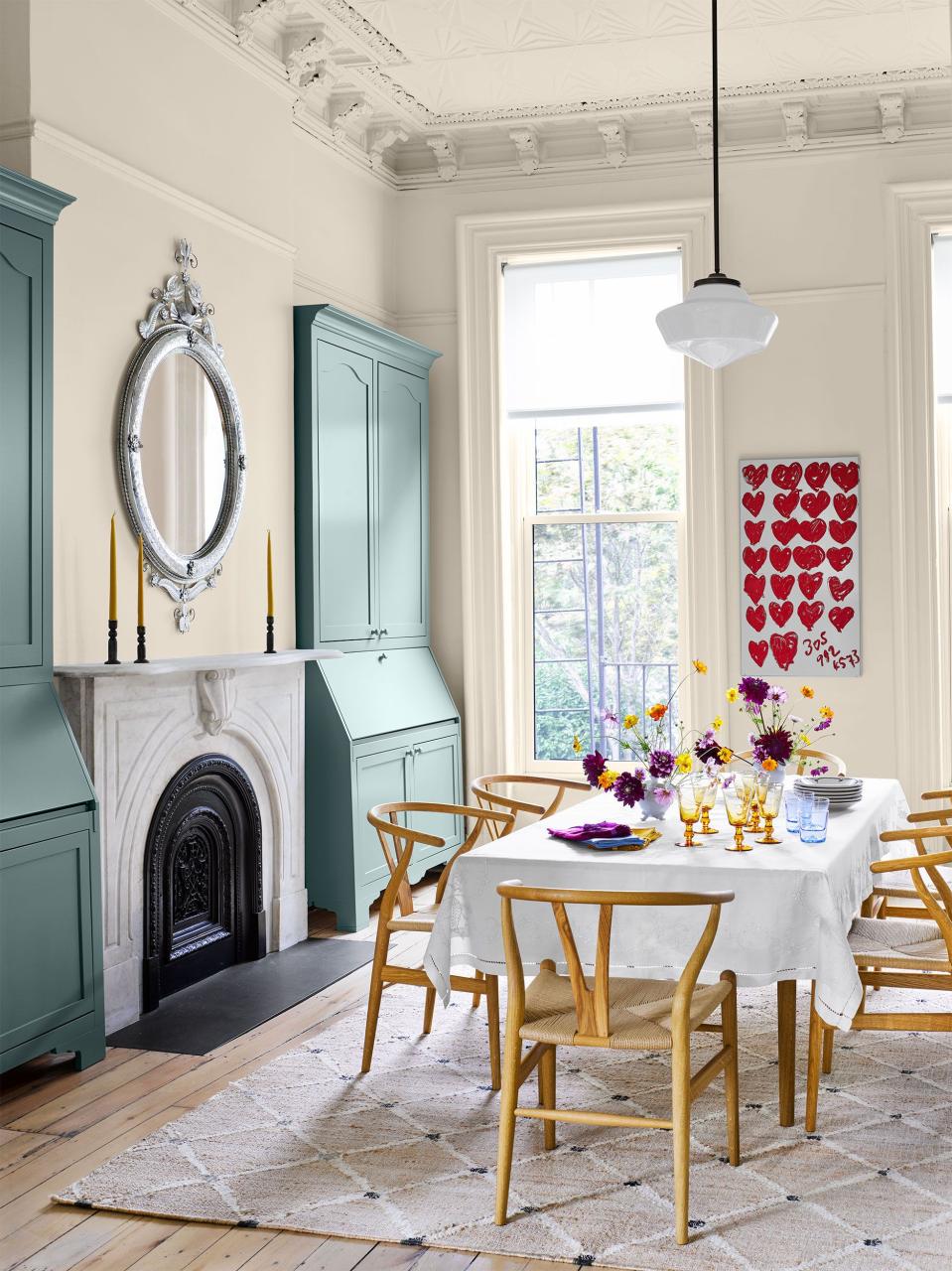 dining room with light wood plank flooring and a long table set with white tablecloth and simple wooden chairs pulled up to it on a hatchwork rug in a neutral beige and flanking the fireplace on the left two tall blue painted secretaries