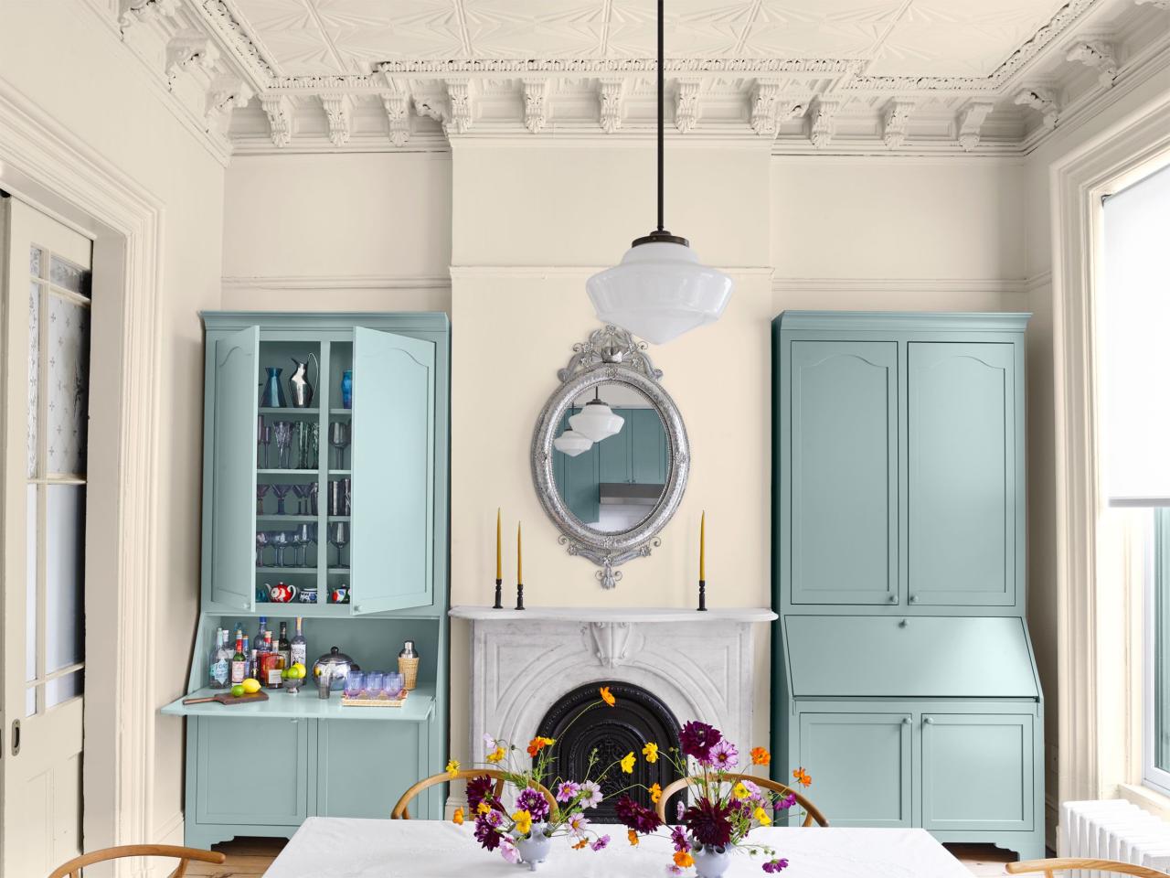 a dining area with off white walls has a table with wooden chairs in foreground, in background is a fireplace with a decorative oval mirror above and is flanked by twin armoires in a seafoam color