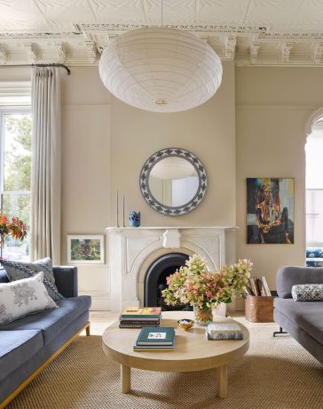 living room has a smoky blue sofa across from a grayish daybed and a light wood oval cocktail table with books and flowers on it, a fireplace with a round mirror flanked by artwork and windows, and large white paper pendant