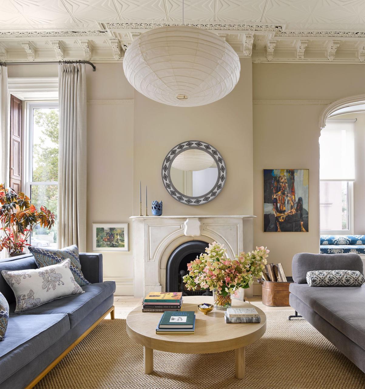 living room has a smoky blue sofa across from a grayish daybed and a light wood oval cocktail table with books and flowers on it, a fireplace with a round mirror flanked by artwork and windows, and large white paper pendant