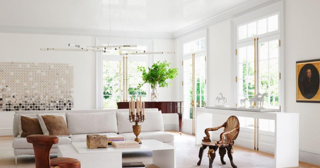 in a white living room is a white sofa, two side chairs, a white cocktail table with books and candelabra, white sideboard, a grand piano, white chandelier with brass arms, a large artwork on back wall, white carpet