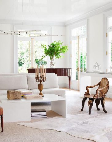 in a white living room is a white sofa, two side chairs, a white cocktail table with books and candelabra, white sideboard, a grand piano, white chandelier with brass arms, a large artwork on back wall, white carpet