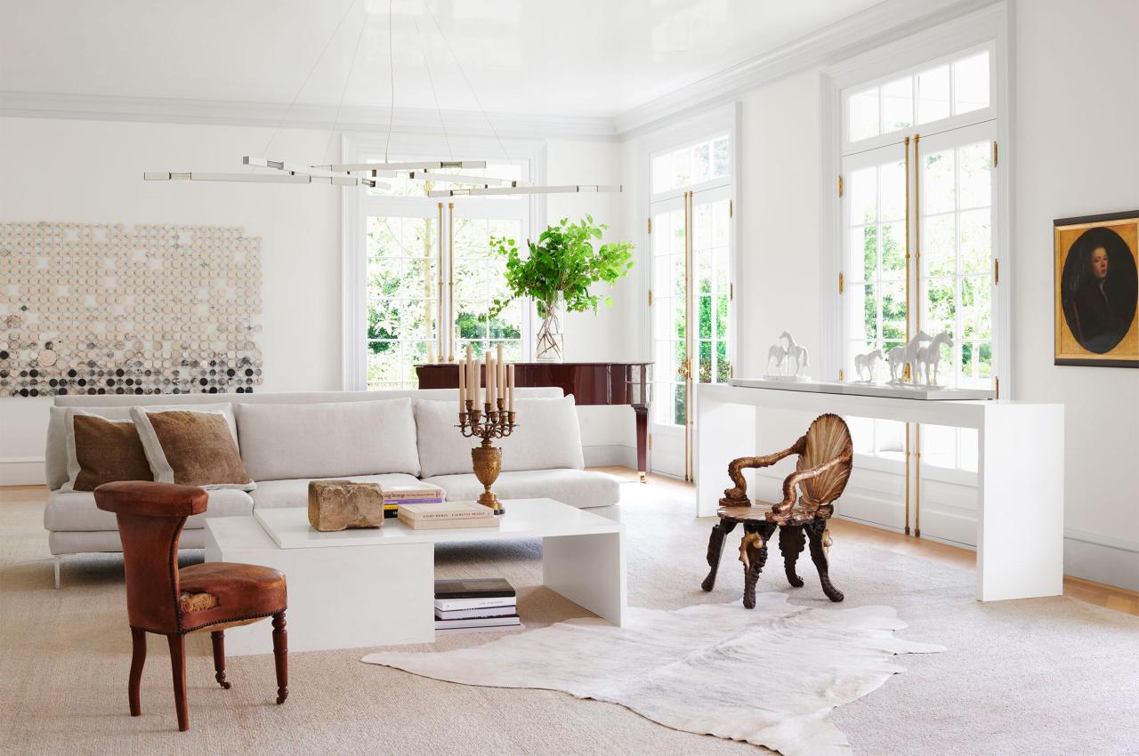 in a white living room is a white sofa, two side chairs, a white cocktail table with books and candelabra, white sideboard, a grand piano, white chandelier with brass arms, a large artwork on back wall, white carpet