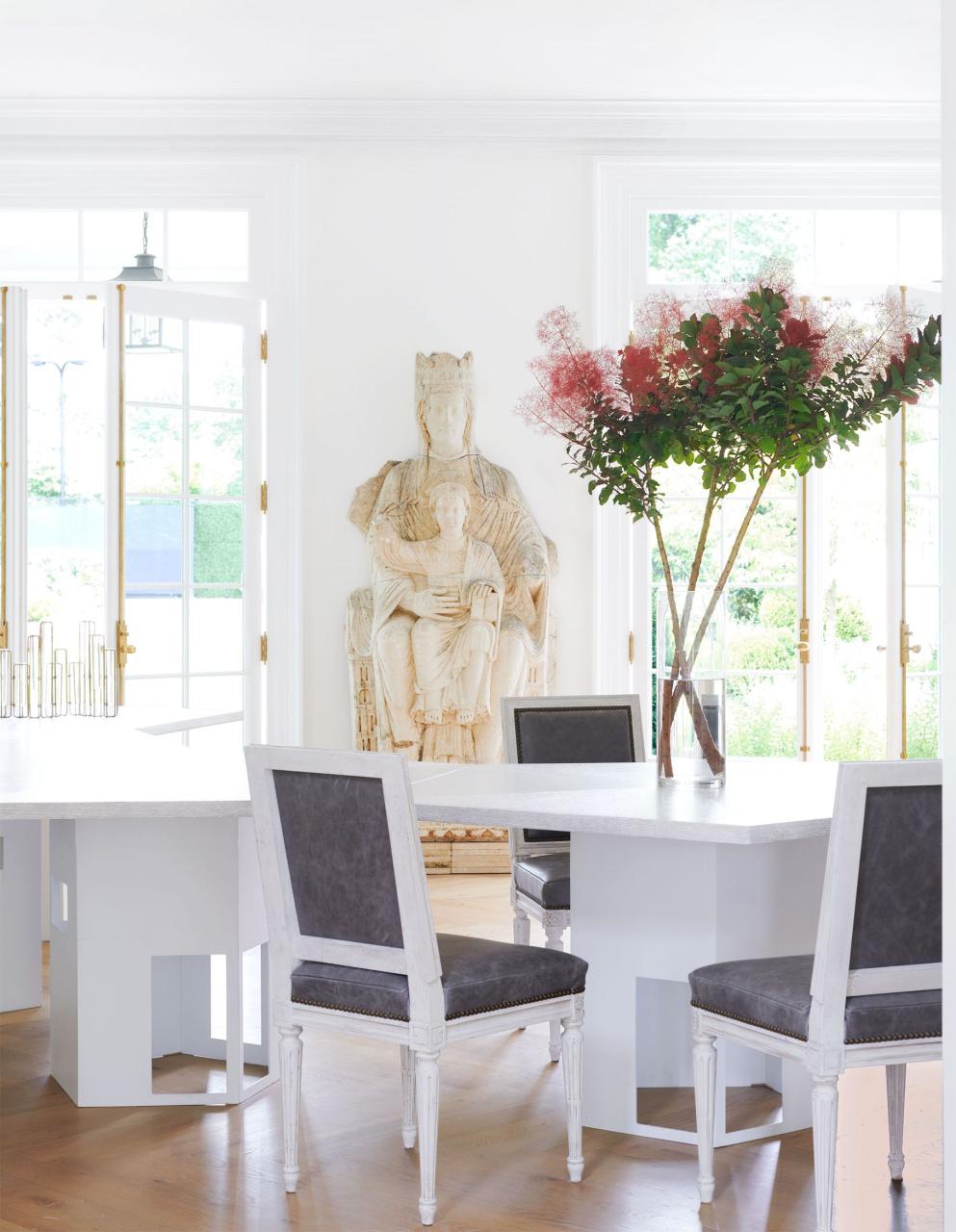 in a white dining room are two glass doors with an antique sculpture of the virgin and child against the wall between them, two geometric shaped white tables, three dining chairs with gray leather seats