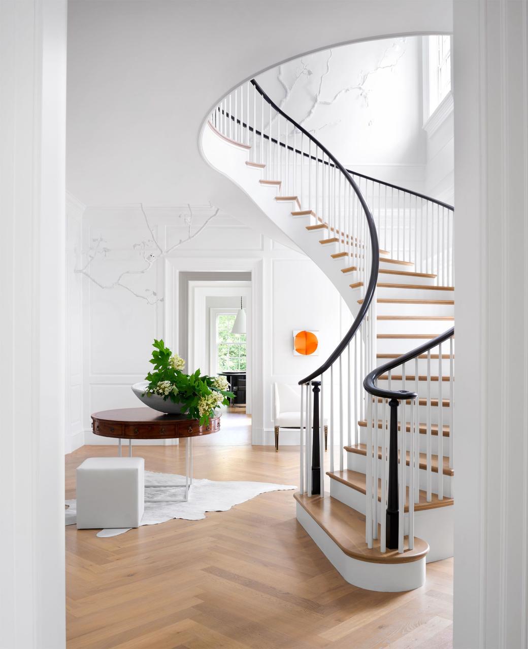 in an entry a grand freestanding curving staircase has natural wood steps, a white balustrade, and black bannisters, wood floor, an antique round table and a white leather cube