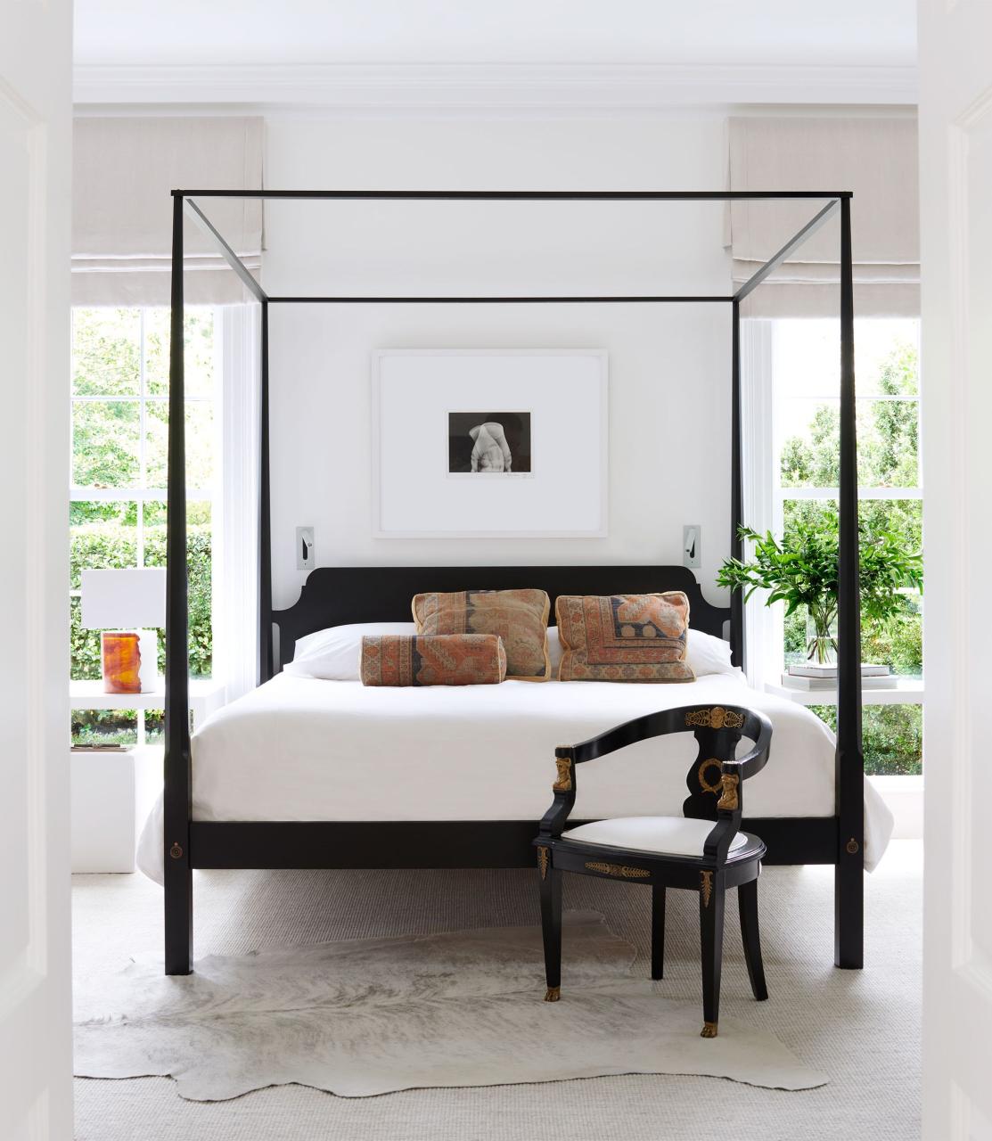 between two windows sits a black four poster bed with white bedding and colorful pillows, white walls, a framed black and white photo, white carpet and a white cow hide rug, and an antique empire chair