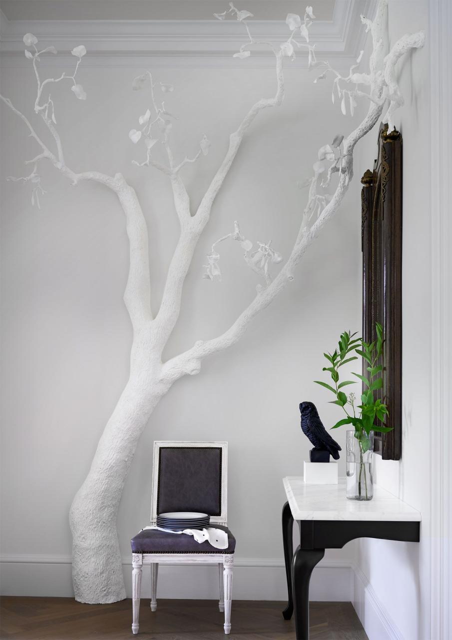 in a corner of room with white walls is a console with curved black legs and a white marble top and a decorative mirror above, a white dining chair, and a floor to ceiling white tree sculpture with branches