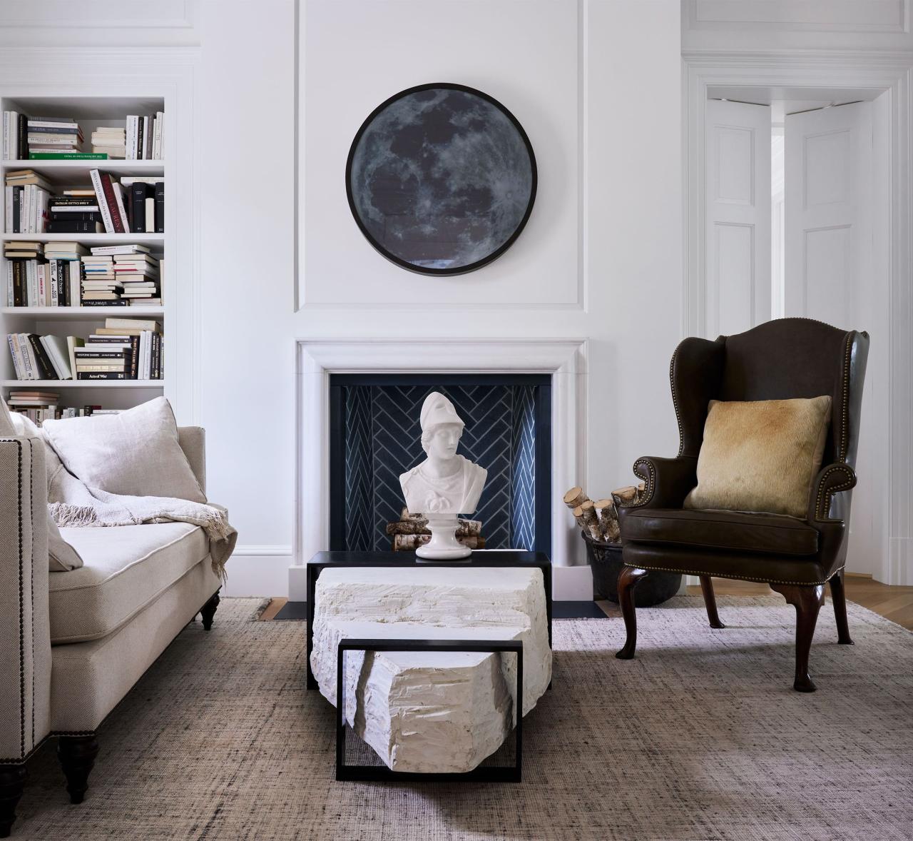 a living room with a white sofa, a cement and black steel cocktail table and a marble bust atop it, a leather wingback chair, a fireplace with a round artwork above, built in bookshelves, and a patterned rug