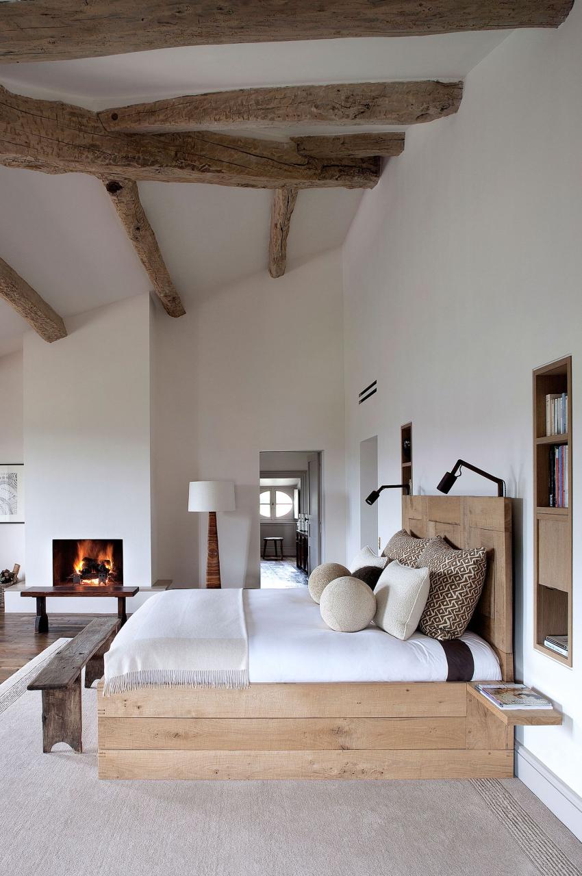 a bedroom with white walls and a few ceilng beams and a fireplace and simple platform bed