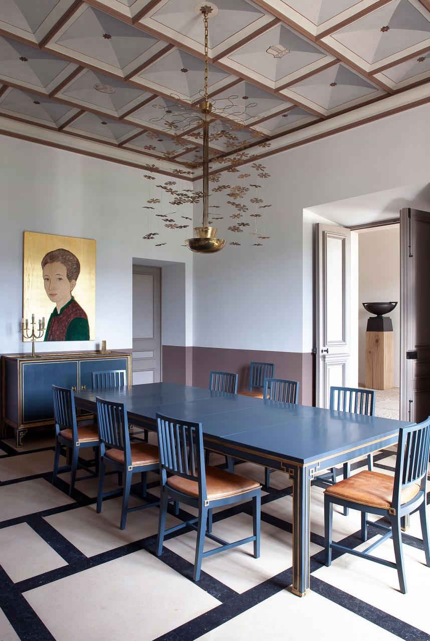 a dining room with blue table and chairs a chandelier overhead and a modern colorful portrait on the wall behind